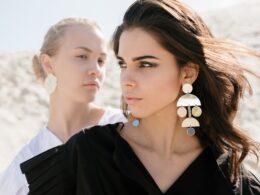 Attractive brunette woman with elegant hairdo wearing black dress with fashionable earrings and makeup standing in front of young blond female friend with simple hairstyle and without makeup in white shirt