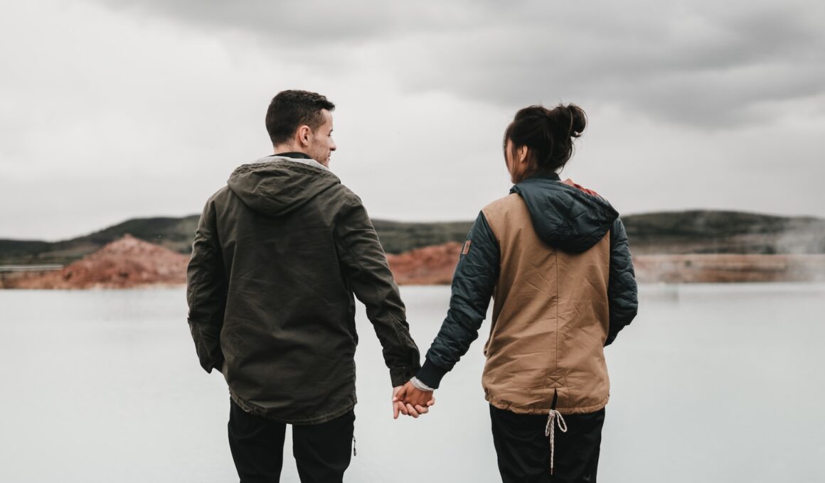 couple holding hand front of body of calm water with mountain distance